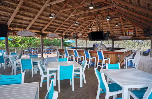 Open-air tiki-style bar at Hilton Cocoa Beach featuring a thatched roof, bamboo bar, and blue-and-white patio seating under ceiling fans and pendant lights.