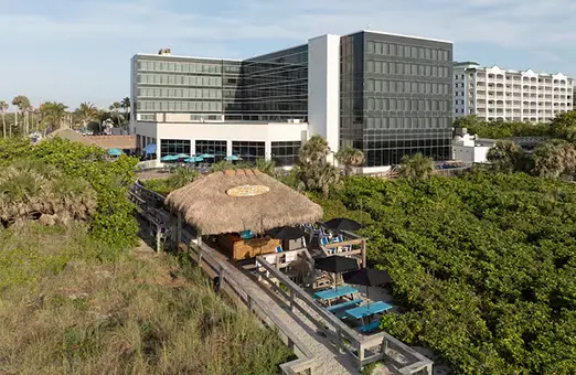 Aerial view of the Hilton Cocoa Beach Oceanfront hotel with a thatched-roof tiki bar and boardwalk seating nestled among coastal greenery.