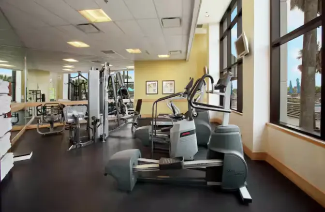Hotel gym at Hilton Cocoa Beach featuring treadmills, ellipticals, weight machines, mirrors, and large windows with outdoor views.