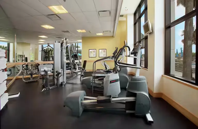 Hotel gym at Hilton Cocoa Beach featuring treadmills, ellipticals, weight machines, mirrors, and large windows with outdoor views.
