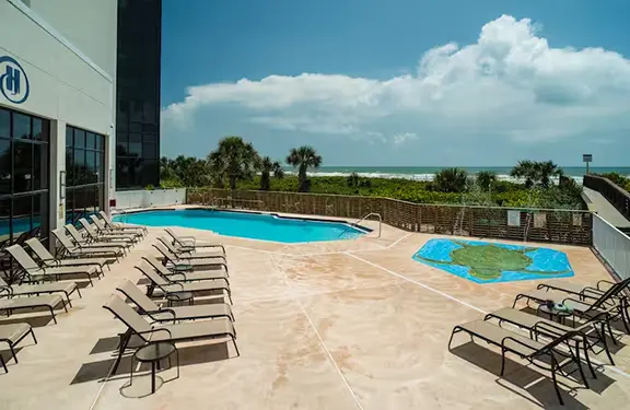 Hilton Cocoa Beach oceanfront pool deck featuring a swimming pool, splash pad, numerous lounge chairs, and views of the Atlantic Ocean.