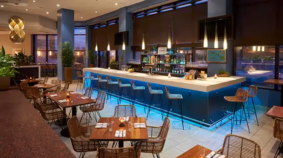 Contemporary bar and lounge area at Hilton Cocoa Beach featuring a long counter with high-top chairs, glowing blue accent lighting, and wooden dining tables with wicker seating.