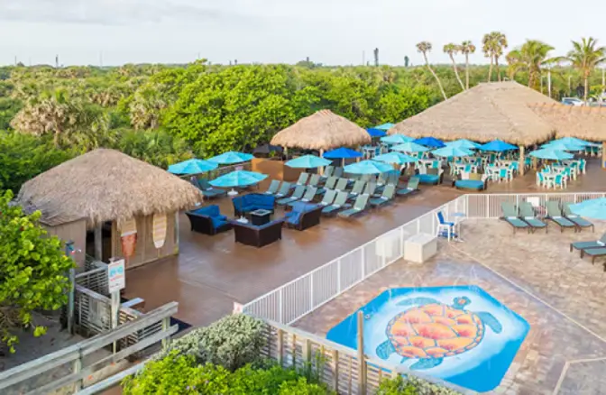 Hilton Cocoa Beach outdoor deck area with thatched-roof cabanas, blue umbrellas, lounge chairs, and a children’s splash pad featuring a large sea turtle graphic.