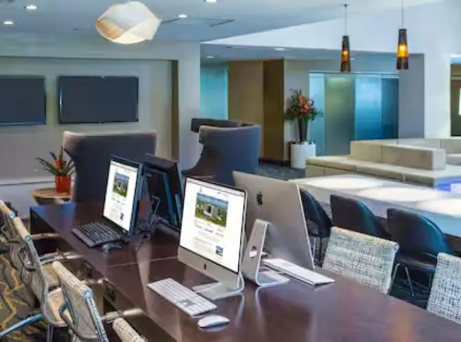 Modern business center at Hilton Cocoa Beach with iMacs and PCs on a long shared desk, surrounded by woven chairs and contemporary decor.