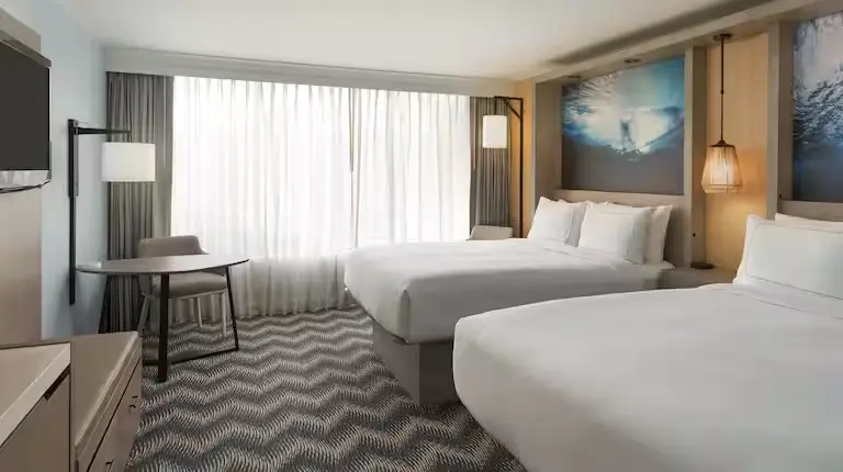 Modern guest room at Hilton Cocoa Beach featuring two queen beds, a work desk with chair, large window with sheer curtains, and ocean-themed wall art.