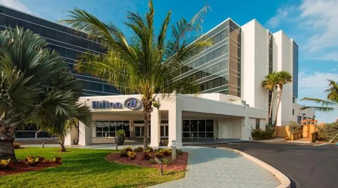 Front entrance of the Hilton Cocoa Beach Oceanfront hotel featuring palm trees, modern architecture, and a curved driveway under clear blue skies.
