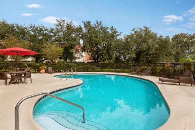 Elegant outdoor pool at Ramada Suites, complete with pristine water, ample lounge chairs, palm trees, and shaded areas under umbrellas, set against the backdrop of the hotel’s contemporary architecture.