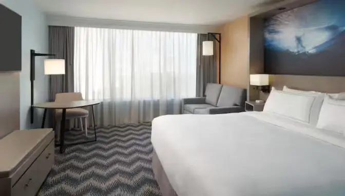 Modern Hilton Cocoa Beach room with one king bed, a gray sofa, a small work desk with chair, and a large window with sheer curtains.