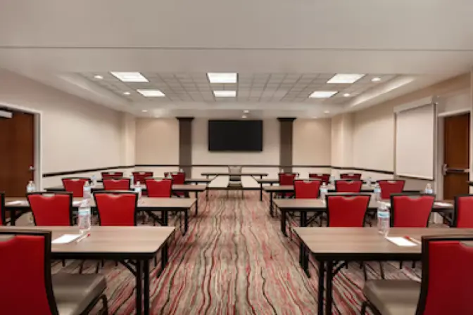 Spacious business center at Ramada Suites featuring a large television at the front of the room with many tables and chairs set up.