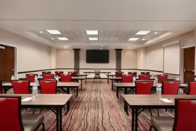 Spacious business center at Ramada Suites featuring a large television at the front of the room with many tables and chairs set up.