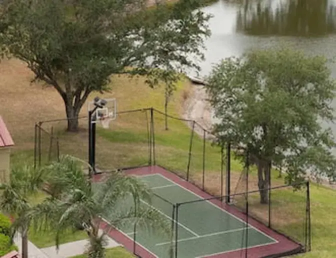 Outdoor sports court with a basketball hoop, net fencing, and green playing surface, situated next to a pond and trees at Residence Inn by Marriott.