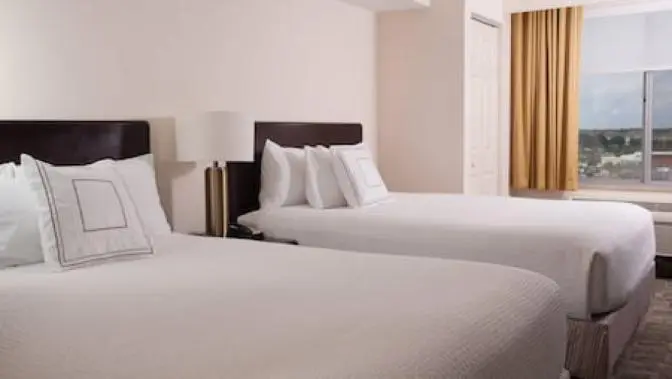 Hotel room at the SpringHill Suites by Marriott featuring two queen beds with white bedding, a modern side table lamp and a large window overlooking a town center.