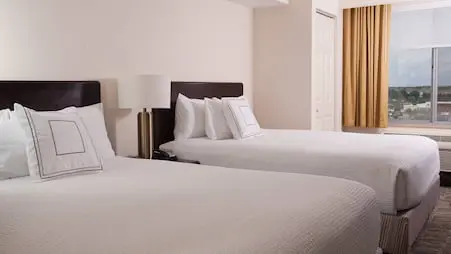 Hotel room at the SpringHill Suites by Marriott featuring two queen beds with white bedding, a modern side table lamp and a large window overlooking a town center.