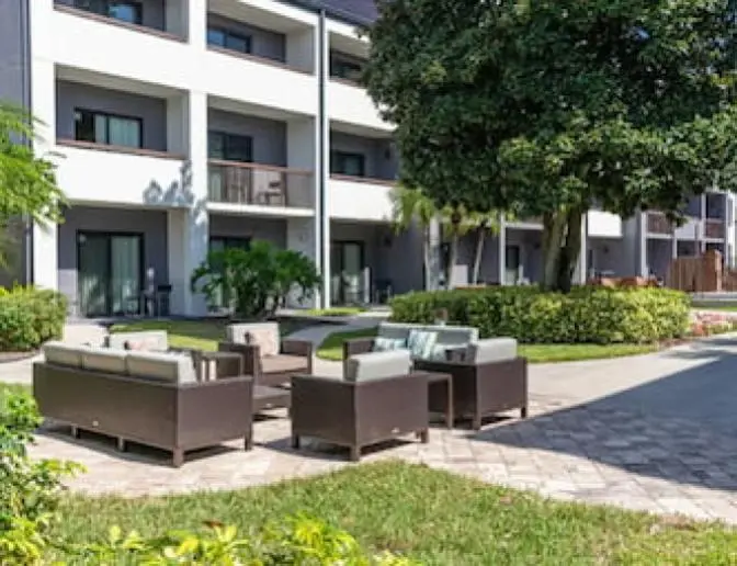 Outdoor patio area at the Courtyard by Marriott hotel featuring comfortable wicker seating with cushions, surrounded by lush greenery and the exterior of the hotel building in the background.