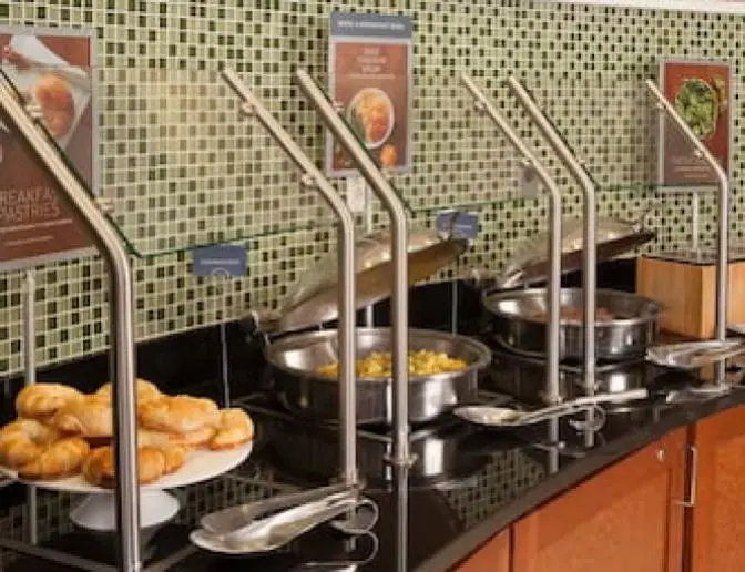 Hot breakfast station at SpringHill Suites by Marriott with trays of pastries, eggs, and sausages, set against a green tile backsplash and displayed under metal serving lids.