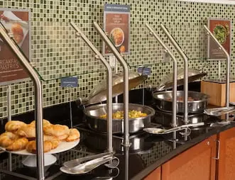 Hot breakfast station at SpringHill Suites by Marriott with trays of pastries, eggs, and sausages, set against a green tile backsplash and displayed under metal serving lids.