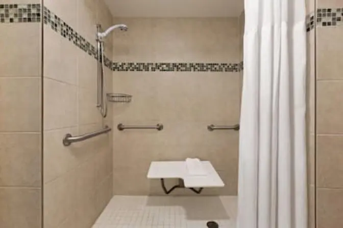 Bathroom inside a suite at the Ramada Suites Orlando International Airport Hotel, featuring an accessible shower with bench.