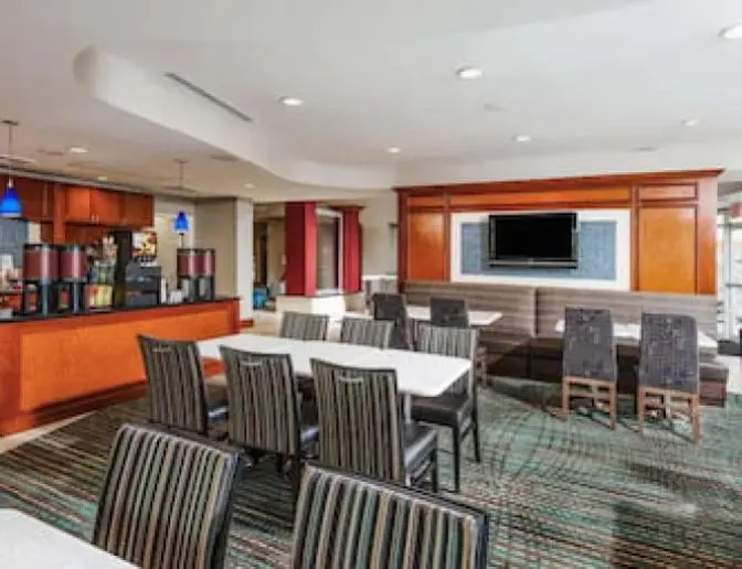 Dining area at Residence Inn by Marriott featuring a communal table with striped chairs, booth seating with a wall-mounted TV, coffee station, and modern decor with blue pendant lights.