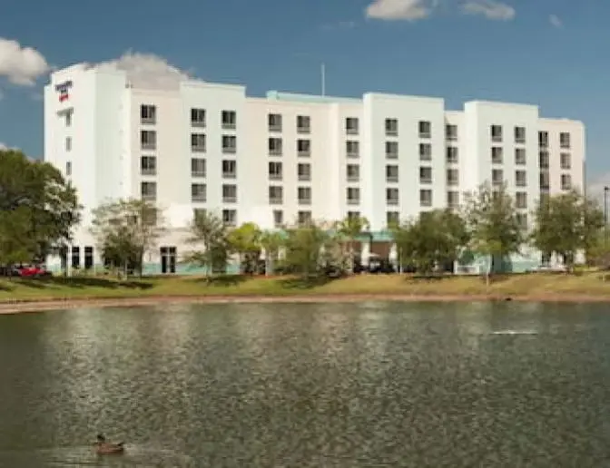 Wide view of SpringHill Suites by Marriott, a multi-story hotel with a white and light blue exterior, seen from across a pond with trees and greenery surrounding the building.