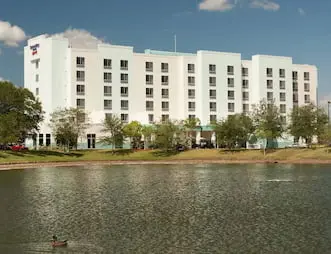 Wide view of SpringHill Suites by Marriott, a multi-story hotel with a white and light blue exterior, seen from across a pond with trees and greenery surrounding the building.