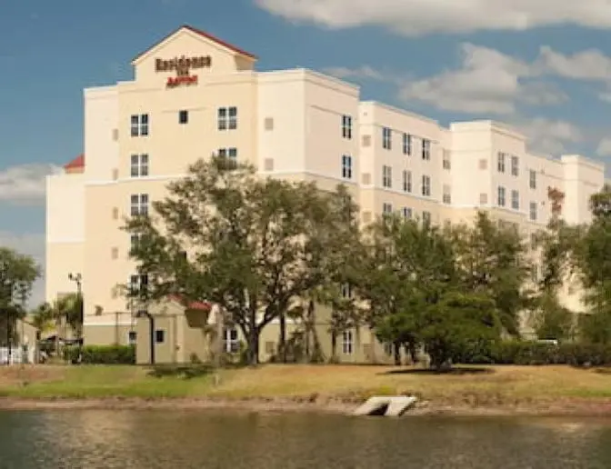 Exterior view of Residence Inn by Marriott with a beige facade, set against a blue sky with trees and a pond in the foreground.