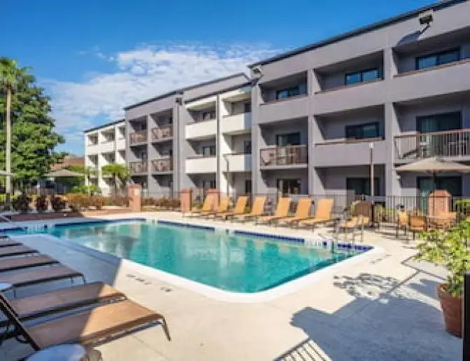 Outdoor swimming pool at the Courtyard by Marriott hotel, featuring lounge chairs and umbrellas around the pool area, with the hotel building and palm trees in the background.