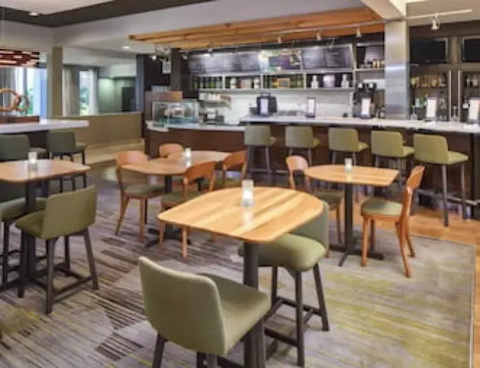 Hotel dining area at the Courtyard by Marriott, showcasing a mix of wooden tables, green chairs, and bar stools, with a bar counter and menu board in the background.