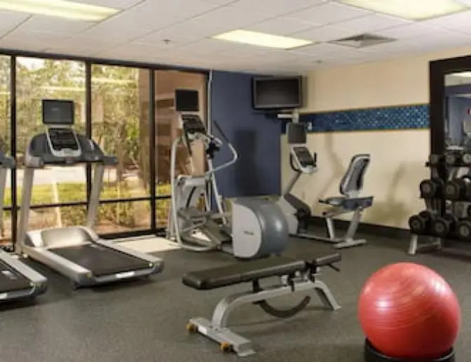 Hampton Inn Orlando International Airport Hotel fitness center with a variety of exercise equipment, including treadmills, an elliptical machine, stationary bike, weight bench, and a rack of dumbbells. The room features large windows with a view of the outside and a wall-mounted TV.