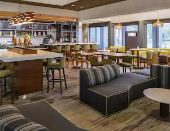 Courtyard by Marriott hotel dining area, highlighting a variety of seating options including high top tables, green chairs, and striped lounge sofas, with a well stocked bar and contemporary lighting fixtures.