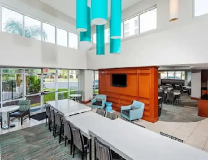 Bright dining area at Residence Inn by Marriott featuring a large communal table with chairs, comfortable seating with teal armchairs, blue pendant lights, and a wall-mounted TV.