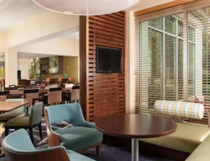Modern dining area in the Hilton Garden Inn Orlando Airport Hotel featuring blue and green seating, wooden tables, and a TV mounted on a wooden divider.