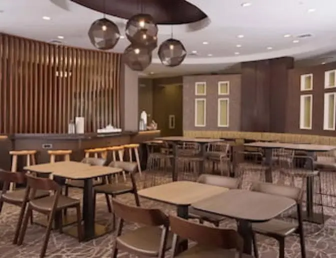 Dining area at SpringHill Suites by Marriott featuring modern wooden tables and chairs, bar-height seating with stools, geometric pendant lights, and a decorative wooden slat partition.