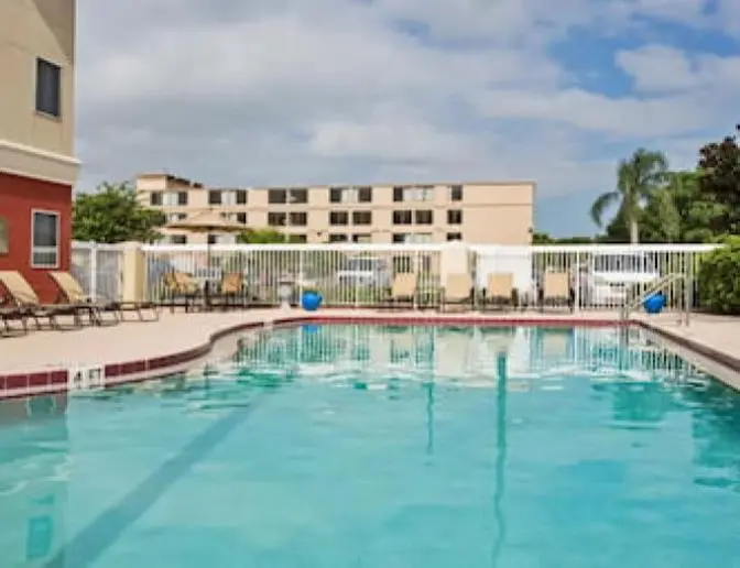 Outdoor swimming pool surrounded by tan lounge chairs with tan umbrellas, under a bright afternoon sky.