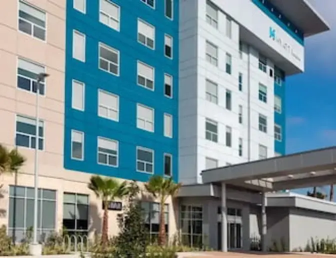Exterior view of Hyatt House Hotel near Orlando International Airport, featuring a modern multi-story building with teal and white accents, a welcoming entrance, and palm trees under a clear blue sky.