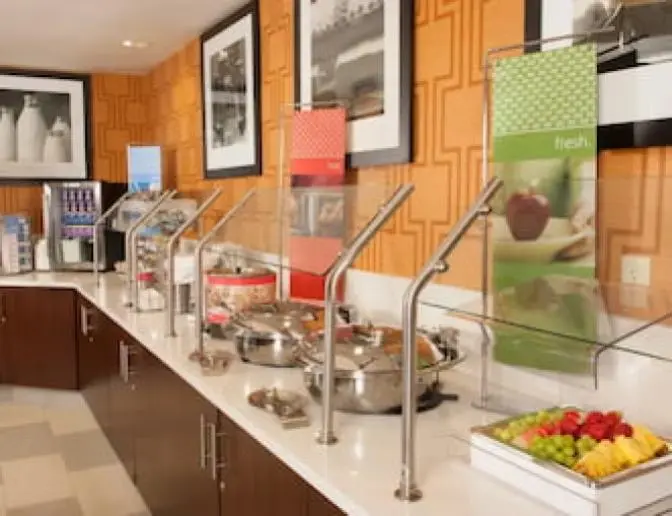 Hampton Inn Orlando International Airport Hotel breakfast buffet counter with a variety of hot and cold food options, including fresh fruit, yogurt, and cereals. The area features stainless steel food warmers, colorful signage, and framed artwork on patterned orange walls.