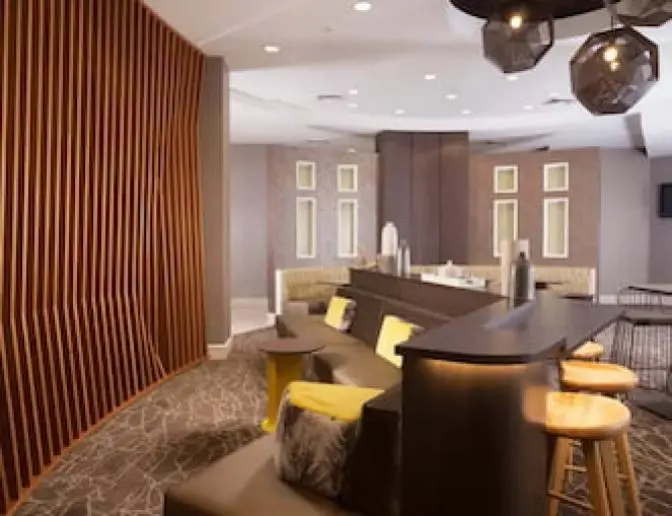 Lobby seating area at SpringHill Suites by Marriott featuring a curved wooden accent wall, modern geometric lighting, bar-height table with stools, and cushioned seating with decorative pillows.