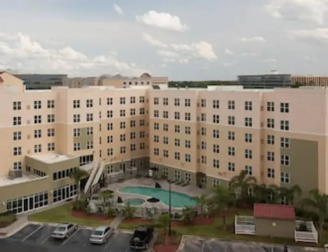 Aerial view of Residence Inn by Marriott featuring a multi-story building with beige and olive green exterior, an outdoor swimming pool, hot tub, and surrounding palm trees.