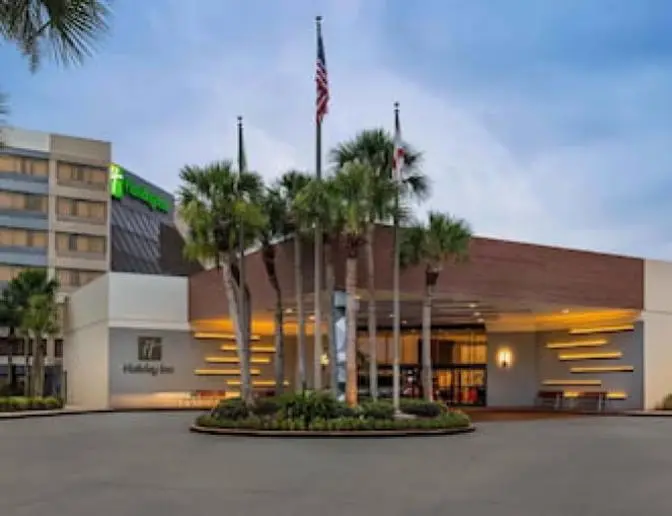 Exterior view of Holiday Inn Orlando International Airport, featuring a modern entrance. with warm lighting, tall palm trees, and flags displayed against an evening sky.