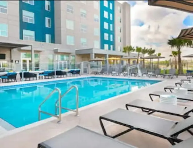 Vibrant outdoor swimming pool surrounded by lounge chairs with black and white striped umbrellas, under a bright sky.