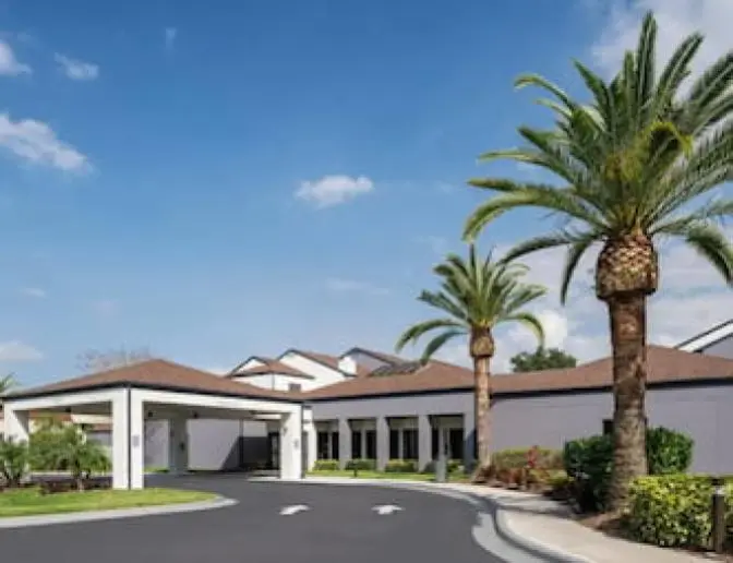 The entrance of the Courtyard by Marriott Orlando International Airport Hotel in the daytime, featuring a portico, palm trees, and a neatly landscaped garden.