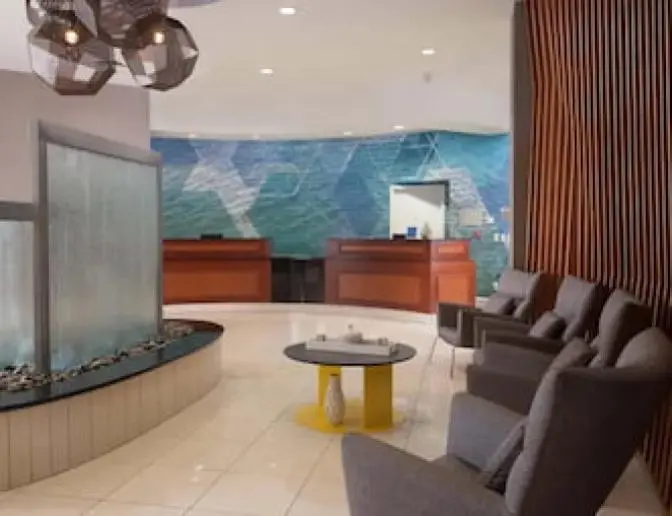Modern lobby of SpringHill Suites by Marriott featuring a reception desk, a water feature with pebbles, comfortable seating area with gray chairs, and contemporary decor including geometric pendant lights.