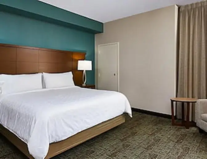 Hotel room with a king-sized bed, wooden headboard, bedside lamps, and a comfortable chair next to a large window.
