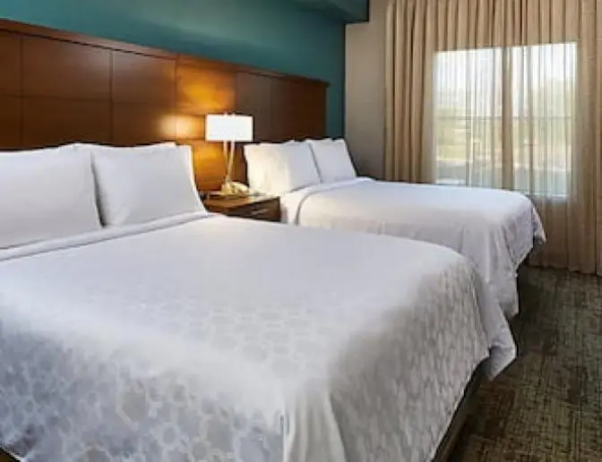 Staybridge Suites room featuring two queen beds, a nightstand with a lamp, and a window with drapes.