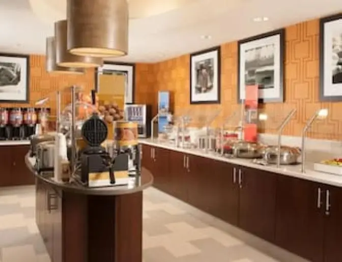 A breakfast buffet area in the Hampton Inn Orlando International Airport Hotel featuring a variety of food options, including a waffle maker, juice dispensers, and hot food stations. The space has warm lighting, dark wood cabinetry, and framed artwork on the orange-patterned walls.