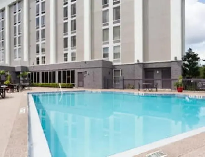 Outdoor swimming pool at Hampton Inn Orlando International Airport Hotel with a surrounding deck area that includes tables and chairs. The pool is adjacent to a tall building with large windows and landscaped with potted plants.