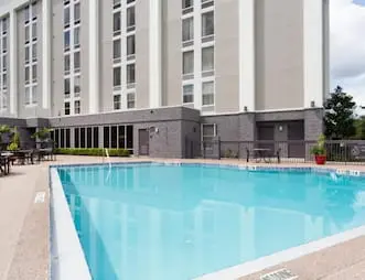 Outdoor swimming pool at Hampton Inn Orlando International Airport Hotel with a surrounding deck area that includes tables and chairs. The pool is adjacent to a tall building with large windows and landscaped with potted plants.