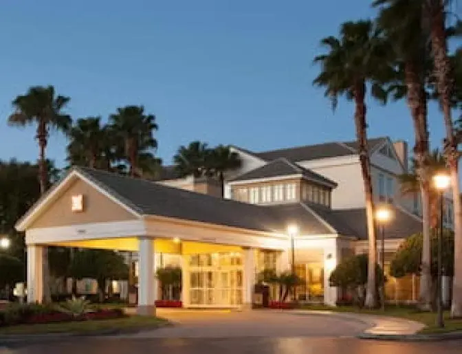Exterior view of Hilton Garden Inn Orlando Airport Hotel entrance at dusk, surrounded by tall palm trees and illuminated by warm lighting.