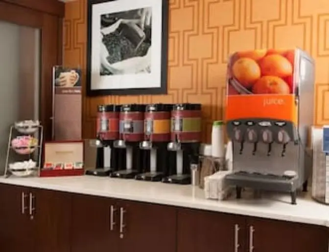 Hampton Inn Orlando International Airport Hotel beverage station featuring coffee dispensers, a juice machine, and a variety of tea options. The counter includes a selection of creamers, sugar, and cups, with framed artwork and an orange-patterned wall in the background.