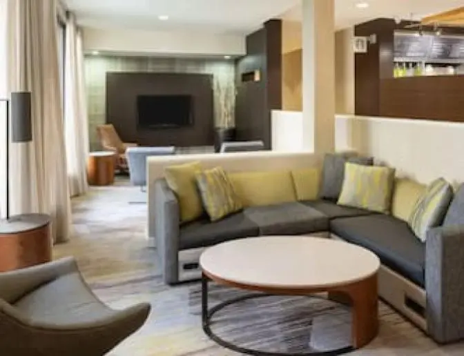 Lobby sitting area at the Courtyard by Marriott, showcasing a stylish sectional sofa with accent pillows, a round coffee table, and a flat-screen TV on a dark wood-paneled wall.