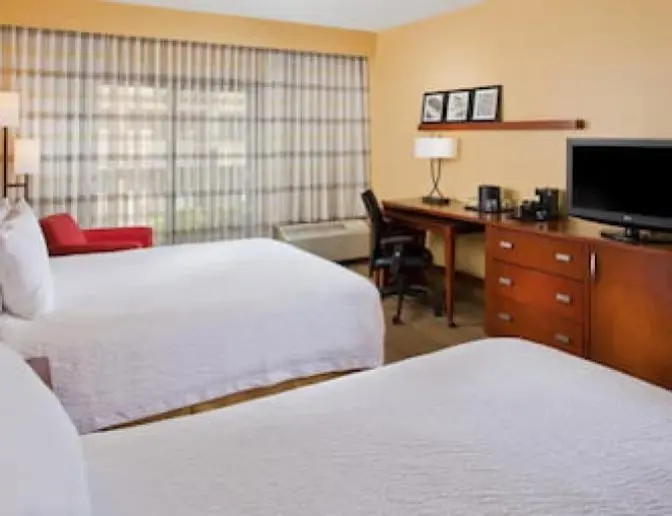 Hotel room at the Courtyard by Marriott featuring two queen beds with white bedding, a work desk with a lamp and chair, a flat-screen TV, and a red armchair near the window.